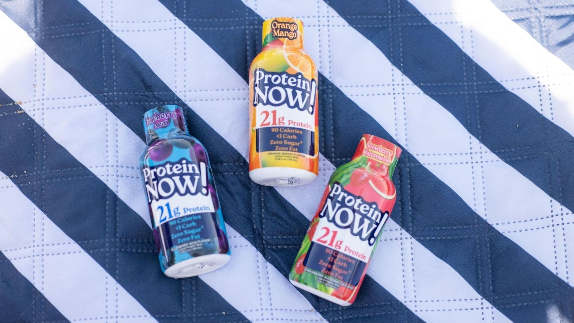 Three colorful Protein Now! 21g protein shot bottles arranged on a blue-and-white patterned blanket.