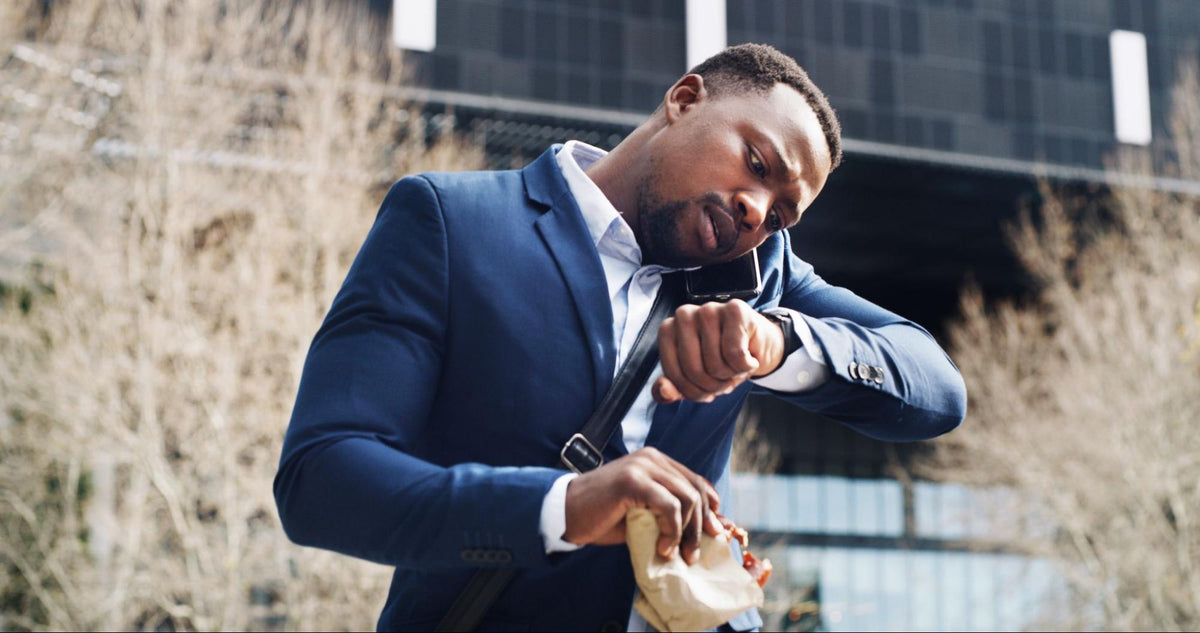 Businessman in a blue suit checking his watch while eating a sandwich outdoors near an office building.