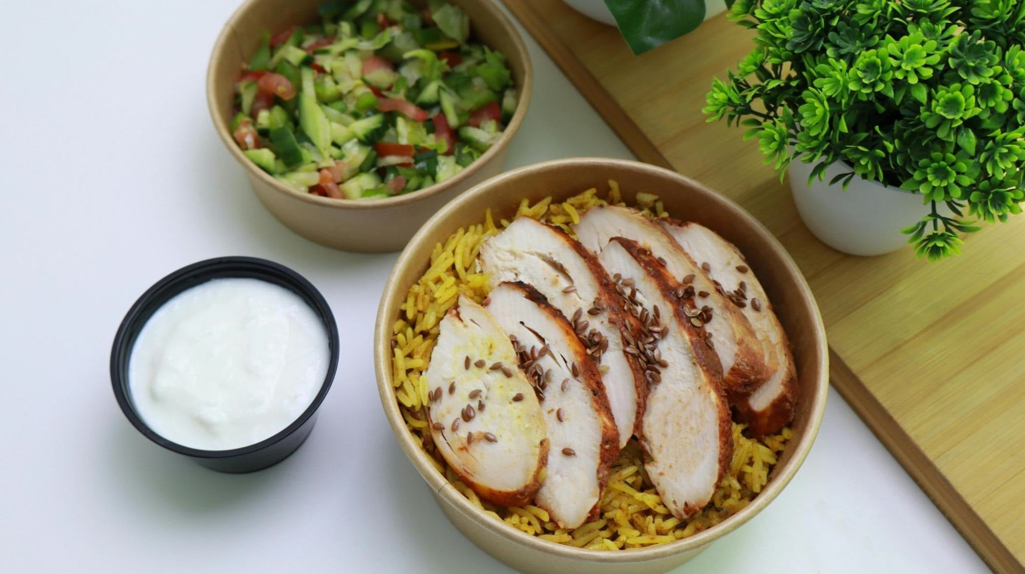 An image of a bowl of sliced chicken and yellow rice, a bowl of chopped greens, and a black ramekin of a white sauce.