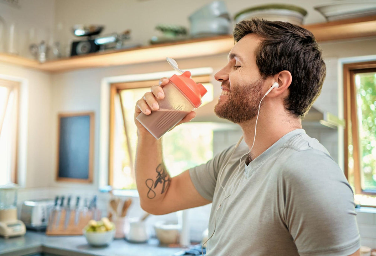 Man enjoying a protein shake while wearing earbuds in a bright home kitchen.