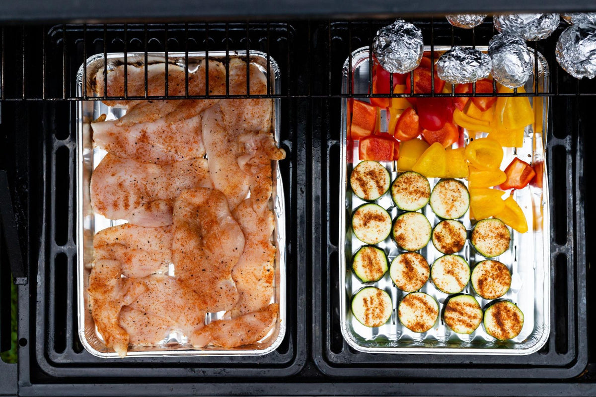 Seasoned chicken breast cutlets, sliced zucchini rounds, chopped red and yellow bell peppers, and foil-wrapped potatoes arranged in aluminum grill trays on a barbecue.