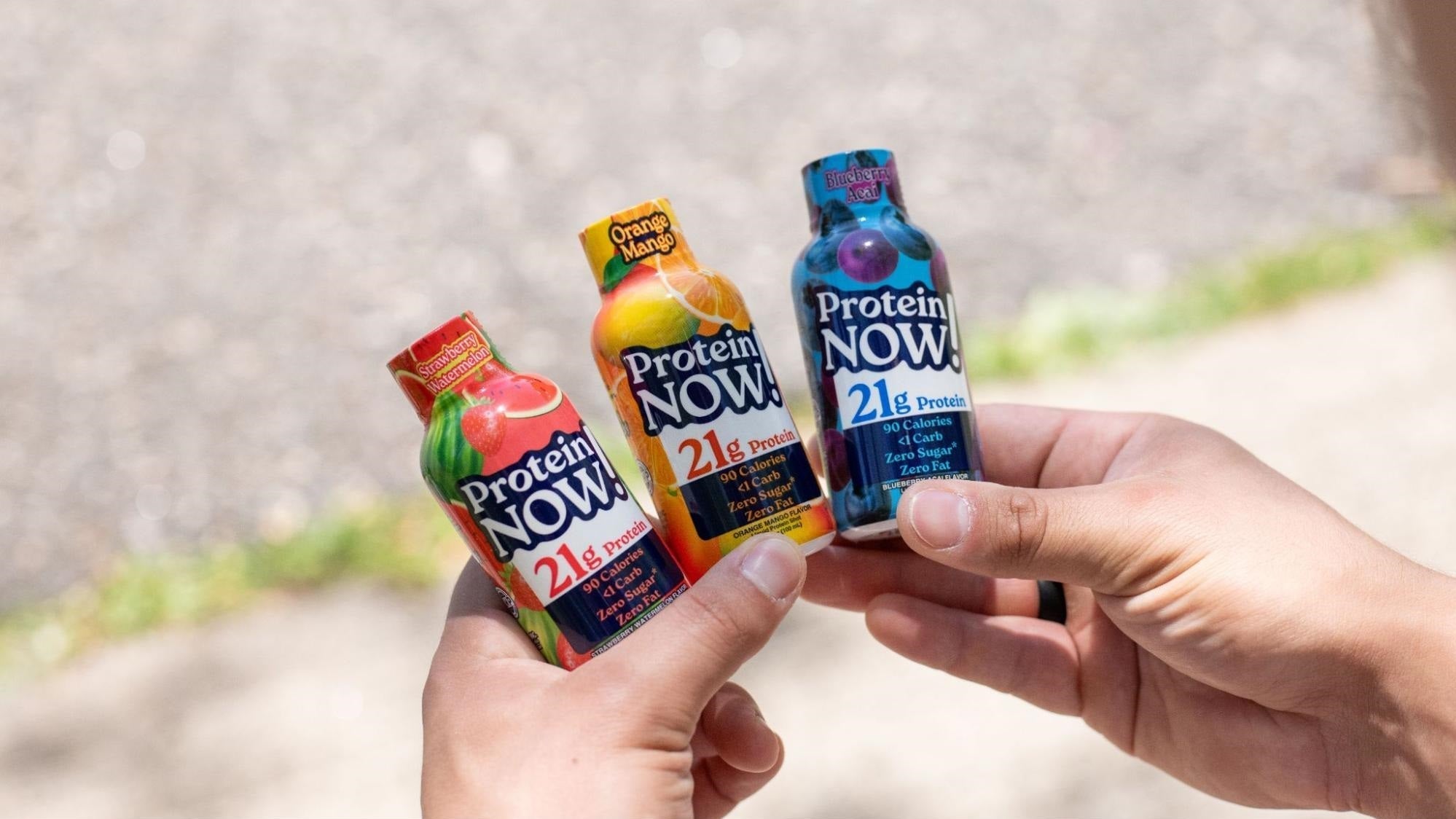 Hands hold three colorful Protein NOW! drinks outdoors, showing red, yellow, and blue bottles labeled with 21 grams of protein.