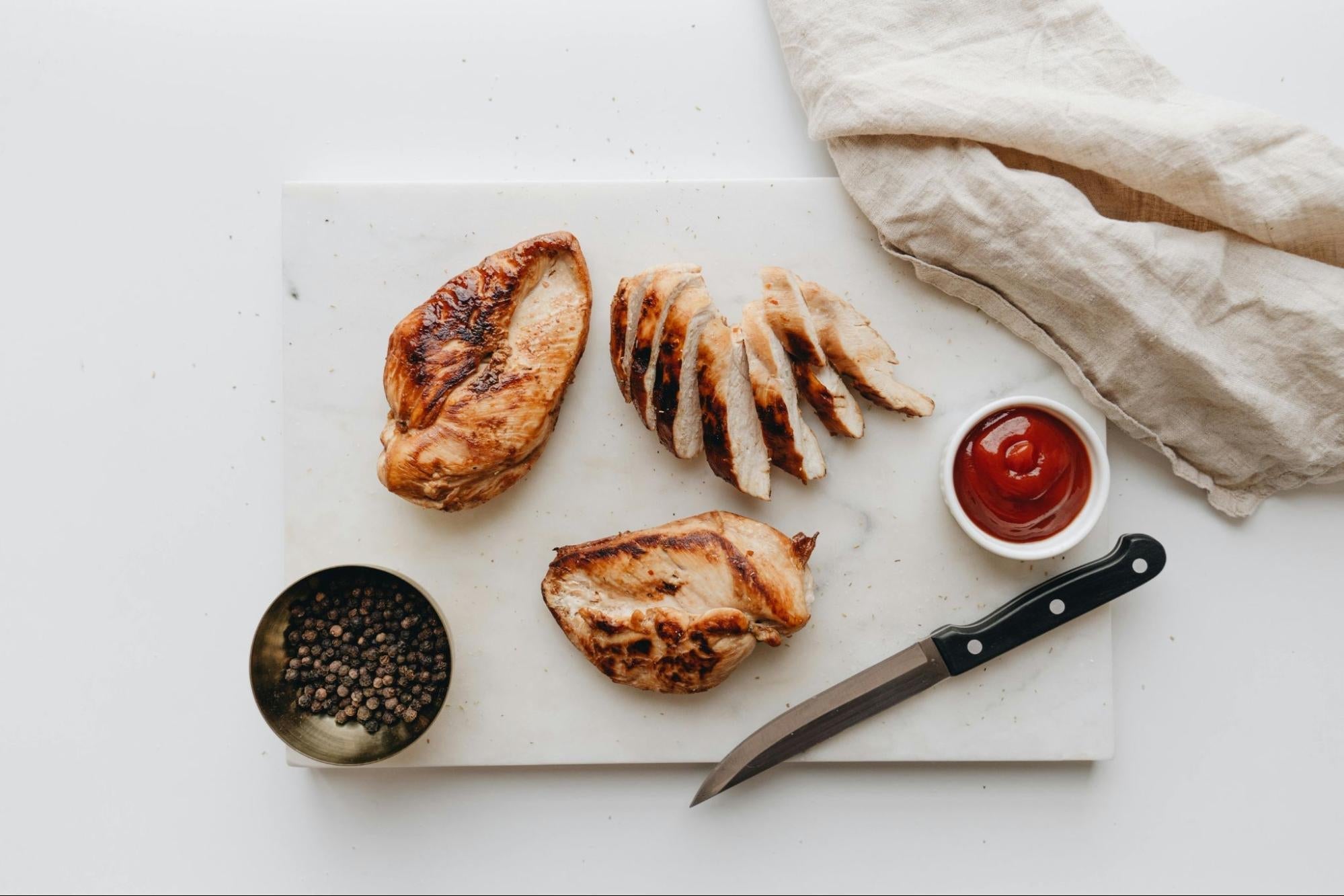 Three pieces of cooked chicken, one sliced, are on a cutting board, along with a steak knife, a dish of peppercorns, and a dish of ketchup, with a beige dish towel on the top right corner.