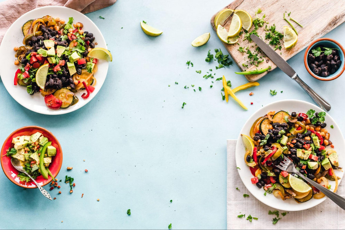 An image of two white ceramic bowls filled with a colorful Mexican-inspired salad containing black beans, diced avocado cubes, roasted zucchini slices, and chopped tomatoes, with lime wedges and chopped cilantro on a wooden cutting board.